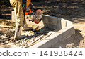 A worker uses a concrete saw to cut a slab on a construction site. The ground is covered with rocks and dust. Tools are nearby, and the work is ongoing 141936424