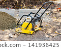 A yellow compactor machine sits on a dirt surface at a construction site. Gravel piles and concrete blocks are nearby. Workers prepare for the next stages 141936425