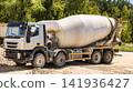 A concrete truck is parked at a construction site. It is surrounded by gravel and trees. Workers can be seen nearby preparing for the next pour 141936427