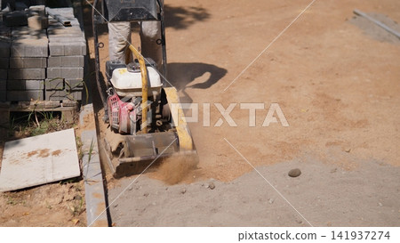 Worker drives vibrating plate compactor across sandy soil beside stacked paving blocks, raising dust while preparing level foundation for driveway, patio, or walkway Worker drives vibrating plate compactor across sandy soil beside stacked paving blocks, raising dust while preparing level foundation for driveway, patio, or walkway 141937274