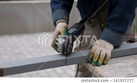 A worker wearing gloves and protective clothing is cutting a metal beam with an angle grinder on a construction site. The scene is focused on the close-up of the worker's hands and tools. A worker wearing gloves and protective clothing is cutting a metal beam with an angle grinder on a construction site. The scene is focused on the close-up of the worker's hands and tools. 141937277