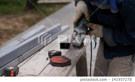 Gloved worker cuts metal tubing with angle grinder while bright sparks scatter beside stacked steel pieces on outdoor workbench, showing hands on fabrication, repair, and construction labor Gloved worker cuts metal tubing with angle grinder while bright sparks scatter beside stacked steel pieces on outdoor workbench, showing hands on fabrication, repair, and construction labor 141937278