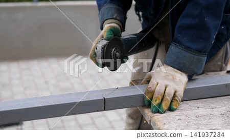 Close view shows worker cutting gray metal beam with angle grinder, wearing protective gloves and work jacket while holding material steady on bench during industrial fabrication and repair task Close view shows worker cutting gray metal beam with angle grinder, wearing protective gloves and work jacket while holding material steady on bench during industrial fabrication and repair task 141937284