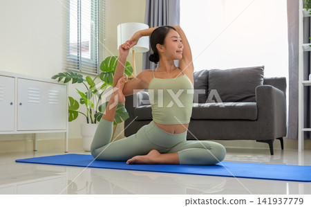 Sporty woman in green top and leggings stretching in Eka Pada Rajakapotasana exercise, One Legged King Pigeon pose on blue mat in bedroom at home. 141937779