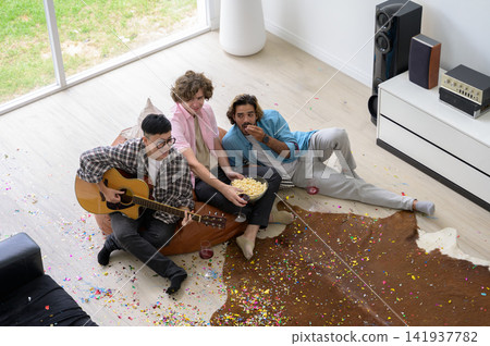 Happy multicultural friends relaxing at home party entertainment together playing acoustic guitar eating popcorn and drinking wine with colorful confetti scattered on floor in modern living room Happy multicultural friends relaxing at home party entertainment together playing acoustic guitar eating popcorn and drinking wine with colorful confetti scattered on floor in modern living room 141937782