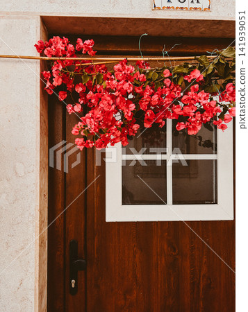 Bright red bougainvillea flowers hang above a dark wooden door with a white framed window, surrounded by a light stone facade Bright red bougainvillea flowers hang above a dark wooden door with a white framed window, surrounded by a light stone facade 141939051