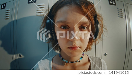 Teenage Girl With Headphones, Staring Directly in Camera, Stands Over School Lockers Teenage Girl With Headphones, Staring Directly in Camera, Stands Over School Lockers 141941015