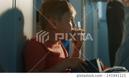 Boy Sitting Comfortably on Floor Near School Lockers, Eating Lunch From Food Container During Recess 141941121