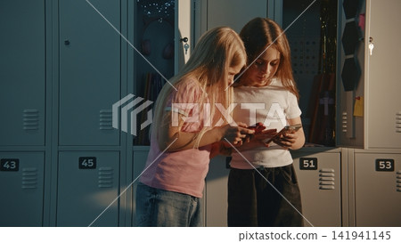 Two Girls by Metal Lockers in Sunlit School Hallway, Both Engrossed in Own Smartphones Two Girls by Metal Lockers in Sunlit School Hallway, Both Engrossed in Own Smartphones 141941145