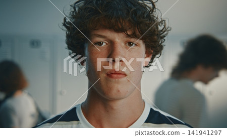 Dark Haired Curly Teenage Boy With Serious Facial Expression, Standing in Bright High School Hallway Dark Haired Curly Teenage Boy With Serious Facial Expression, Standing in Bright High School Hallway 141941297