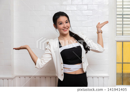 Smiling beautiful Asian woman with both her empty palms extended sideways presenting copy-space on white wall. Smiling beautiful Asian woman with both her empty palms extended sideways presenting copy-space on white wall. 141941405