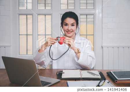 Professional female medical doctor holding a stethoscope check up on a red heart ball in office in the hospital. Concept of health care and medical. Professional female medical doctor holding a stethoscope check up on a red heart ball in office in the hospital. Concept of health care and medical. 141941406