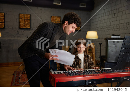 Enthusiastic young girl practicing piano alongside her mentor in cozy studio setting Enthusiastic young girl practicing piano alongside her mentor in cozy studio setting 141942045