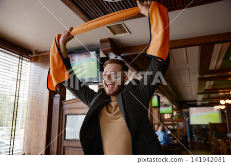 Fan cheers during live game. Enthusiastic spectator celebrates goal amid lively pub setting Fan cheers during live game. Enthusiastic spectator celebrates goal amid lively pub setting 141942081