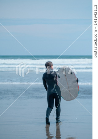 A female surfer walks along the waterline, carrying her surfboard, from behind | A quiet morning on a cloudy beach A female surfer walks along the waterline, carrying her surfboard, from behind | A quiet morning on a cloudy beach 141942112