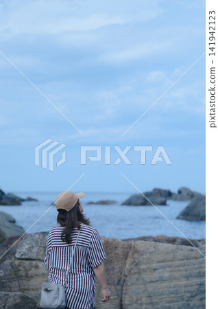 A woman's back as she stands on a rocky shore, gazing at the sea dotted with small islands. A woman's back as she stands on a rocky shore, gazing at the sea dotted with small islands. 141942123