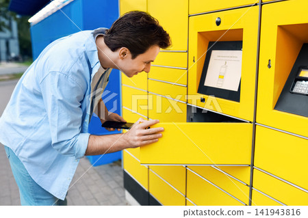 Man using a smartphone for contactless parcel pickup at a yellow self-service terminal. Modern e-commerce delivery service and automated logistics locker. 141943816