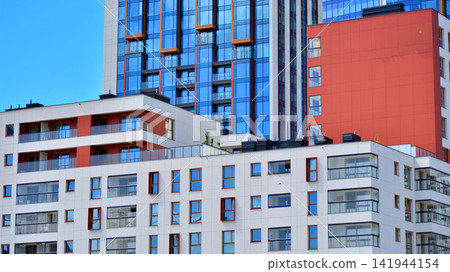 A low-rise residential building stands against a backdrop of tall glass buildings. An urban quarter of modern architecture. A low-rise residential building stands against a backdrop of tall glass buildings. An urban quarter of modern architecture. 141944154