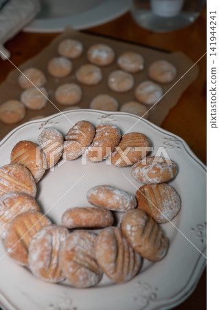 Freshly baked almond cookies dusted with powdered sugar cool on baking paper with another batch served on plate in foreground. Home baking, dessert, recipe, confectionery, food preparation, comfort Freshly baked almond cookies dusted with powdered sugar cool on baking paper with another batch served on plate in foreground. Home baking, dessert, recipe, confectionery, food preparation, comfort 141944241