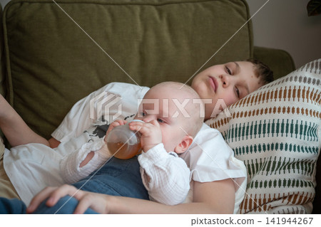 Older brother watches over baby sibling while younger child drinks from bottle resting in his arms on sofa at home. Sibling care, brotherhood, caregiving, responsibility, family bond, childhood 141944267