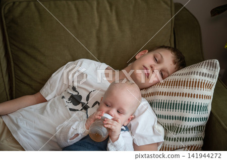 Older brother watches over baby sibling while younger child drinks from bottle resting in his arms on sofa at home. Sibling care, brotherhood, caregiving, responsibility, family bond, childhood Older brother watches over baby sibling while younger child drinks from bottle resting in his arms on sofa at home. Sibling care, brotherhood, caregiving, responsibility, family bond, childhood 141944272