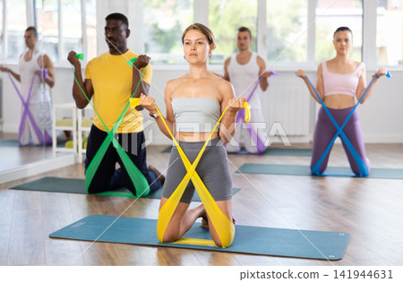 Young woman performs exercises with resistance band during multinational Pilates training, kneeling Young woman performs exercises with resistance band during multinational Pilates training, kneeling 141944631