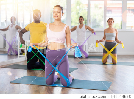 Young woman performs exercises with resistance band during multinational Pilates training, kneeling Young woman performs exercises with resistance band during multinational Pilates training, kneeling 141944638