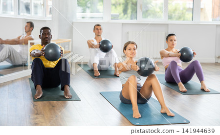 Young woman with multinational athletes group hold fitball in lap knee , raise and lower torso Young woman with multinational athletes group hold fitball in lap knee , raise and lower torso 141944639