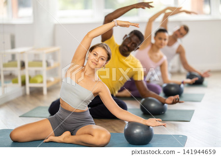 Girl performing side bend with exercise ball during group Pilates session Girl performing side bend with exercise ball during group Pilates session 141944659