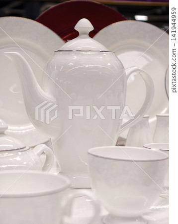 White porcelain cups on saucers tall minimalist teapot and plates behind arranged as premium kitchenware set in refined retail presentation for store shelf display White porcelain cups on saucers tall minimalist teapot and plates behind arranged as premium kitchenware set in refined retail presentation for store shelf display 141944959