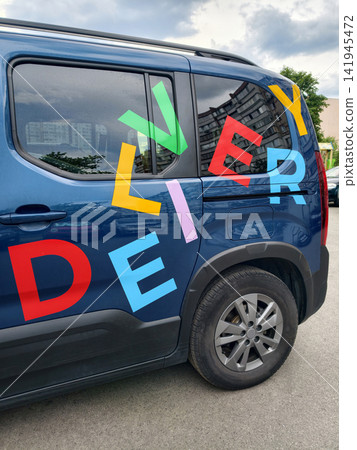 Blue passenger car with large colorful DELIVERY lettering parked in a residential urban neighborhood presenting a clear transportation and logistics concept with everyday city atmosphere Blue passenger car with large colorful DELIVERY lettering parked in a residential urban neighborhood presenting a clear transportation and logistics concept with everyday city atmosphere 141945472