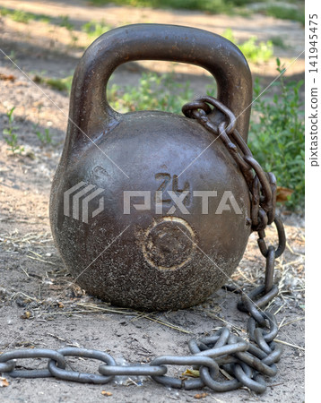 Iron kettlebell weighing 24 kg standing on the ground and secured with a metal chain at an outdoor sports area for strength training and fitness exercises Iron kettlebell weighing 24 kg standing on the ground and secured with a metal chain at an outdoor sports area for strength training and fitness exercises 141945475