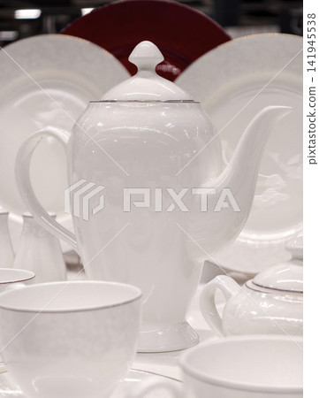 White cups on saucers tall graceful porcelain teapot and plates behind forming elegant kitchenware set in refined retail display concept for luxury tableware marketing White cups on saucers tall graceful porcelain teapot and plates behind forming elegant kitchenware set in refined retail display concept for luxury tableware marketing 141945538