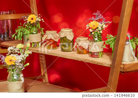 Jars filled with pickles and flowers are placed on a wooden shelf against a bright red background at a market event Jars filled with pickles and flowers are placed on a wooden shelf against a bright red background at a market event 141946610