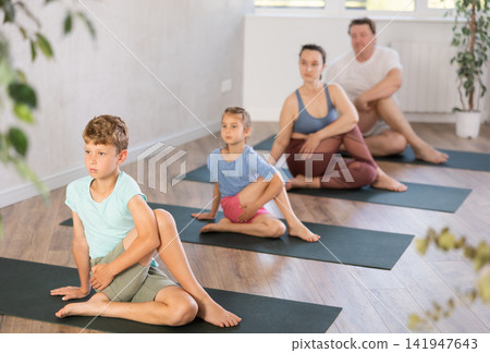 Diligent preteen boy practicing half lord of the fishes pose of yoga with his family Diligent preteen boy practicing half lord of the fishes pose of yoga with his family 141947643