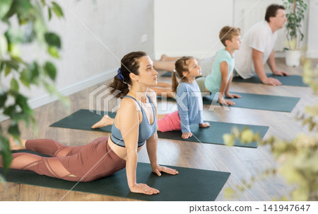 Willing middle-aged woman practicing cobra pose of yoga with her family Willing middle-aged woman practicing cobra pose of yoga with her family 141947647
