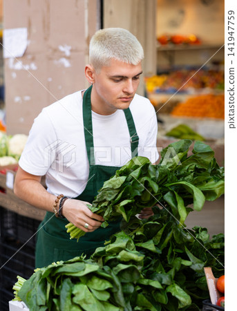 Young guy seller puts spinach in vegetable shop Young guy seller puts spinach in vegetable shop 141947759