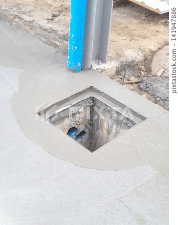 Freshly poured wet concrete floor at a construction site featuring a square drain opening, a blue utility pipe, and a grey structural steel beam. Industrial infrastructure development process. Freshly poured wet concrete floor at a construction site featuring a square drain opening, a blue utility pipe, and a grey structural steel beam. Industrial infrastructure development process. 141947886