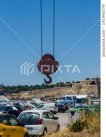 Rusty industrial crane hook suspended on steel cables above a marina parking area, with ancient fortification walls and yachts in Malta.. High quality photo Rusty industrial crane hook suspended on steel cables above a marina parking area, with ancient fortification walls and yachts in Malta.. High quality photo 141948379