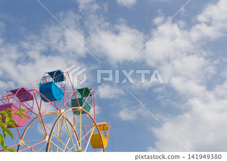A colorful Ferris wheel stands out against the blue sky and clouds, amusement park attractions and an open, expansive view of the sky. A colorful Ferris wheel stands out against the blue sky and clouds, amusement park attractions and an open, expansive view of the sky. 141949380