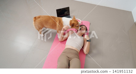Cheerful Asian woman lying on exercise mat laughing while Corgi dog licks her face at home Cheerful Asian woman lying on exercise mat laughing while Corgi dog licks her face at home 141951132