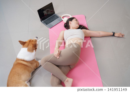 High angle view of Asian woman doing spinal twist yoga stretch near corgi dog High angle view of Asian woman doing spinal twist yoga stretch near corgi dog 141951149