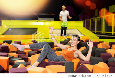 Fitness, fun, leisure and sport activity concept - happy woman fell on soft, safe filler cubes after jumping on trampoline Fitness, fun, leisure and sport activity concept - happy woman fell on soft, safe filler cubes after jumping on trampoline 141951546