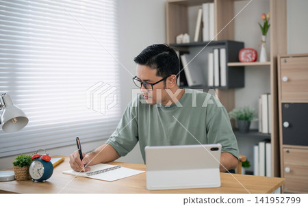 Young man wearing glasses writing notes with pen while using tablet computer at home office desk surrounded by modern decor and natural light window Young man wearing glasses writing notes with pen while using tablet computer at home office desk surrounded by modern decor and natural light window 141952799