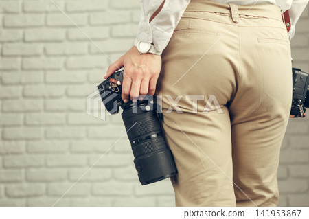 Person holds a camera with a large lens while standing near a brick wall indoors Person holds a camera with a large lens while standing near a brick wall indoors 141953867