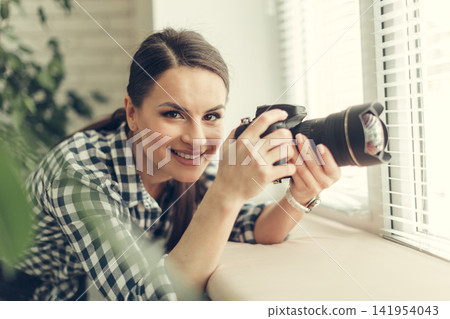 Smiling woman holding camera near window during daytime Smiling woman holding camera near window during daytime 141954043