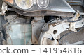 Close-up of a disassembled car front showing a headlight, metal frame brackets, dusty wiring harnesses, translucent washer fluid reservoir, and the radiator. Suggesting collision repair, inspection. 141954816