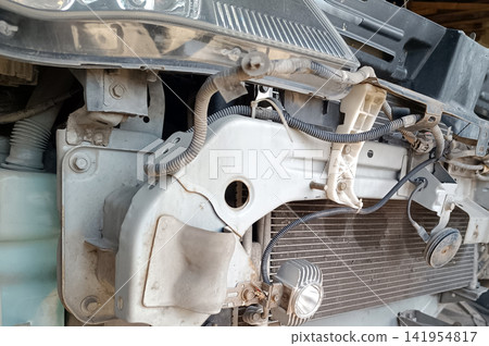 Detailed close-up shot revealing the exposed front structure of a car. The bumper cover is removed, showing the frame rail, radiator, wiring, brackets, bolts, small horn, an LED work light accessory. Detailed close-up shot revealing the exposed front structure of a car. The bumper cover is removed, showing the frame rail, radiator, wiring, brackets, bolts, small horn, an LED work light accessory. 141954817