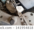 A close-up photograph of a dirty car engine compartment. The central focus is the dust-covered white plastic cap of the windshield washer fluid reservoir, clearly marked  141954818