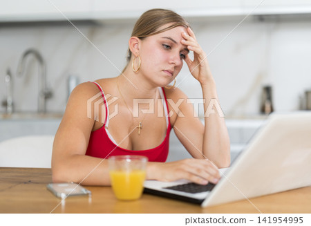Teenage girl having problems with laptop while sitting at table in kitchen Teenage girl having problems with laptop while sitting at table in kitchen 141954995